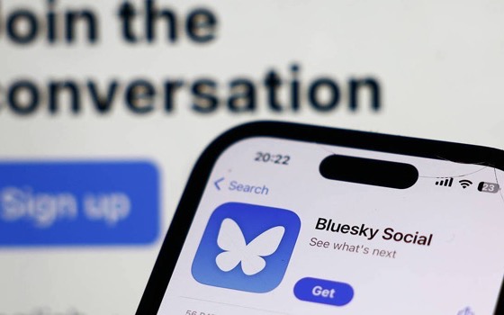 A phone showing the Bluesky Social app listing, in front of a screen reading “Join the conversation”.