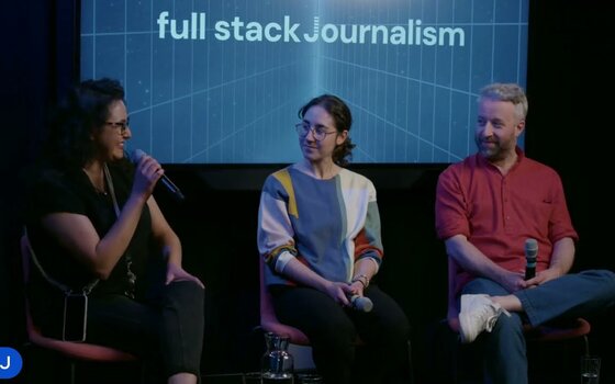 Three people seated on stage with microphones in front of a screen reading “full stack Journalism”.