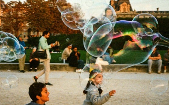 Children chasing giant soap bubbles in a Paris park at dusk.